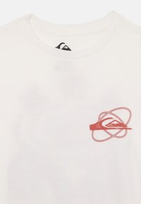T-shirt in cotone bianco con un design grafico rosso che presenta una montagna e elementi circolari sul lato sinistro del petto. Collo rotondo, maniche corte.