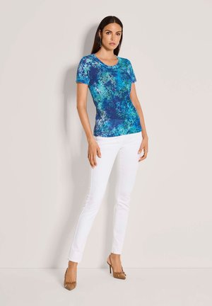 Vrouw staat tegen een muur, draagt een blauw patroon kort mouwtopje, witte strakke broek en bruine hoge hakken.