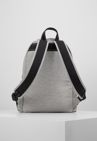 Šedý batoh s hladkým povrchem, černými popruhy a kontrastním prošíváním. Obsahuje horní držadlo a nastavitelné popruhy.