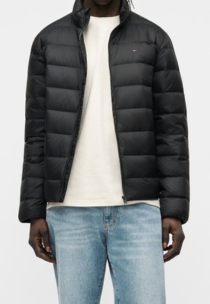 Mężczyzna w czarnej pikowanej kurtce zapinanej na zamek, na białej koszuli i jasnoniebieskich dżinsach, stojący na jednolitym tle.