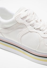 Baskets blanches à semelle épaisse ornées de rayures pastel jaunes, vertes, roses et noires, avec des lacets blancs noués en nœud.