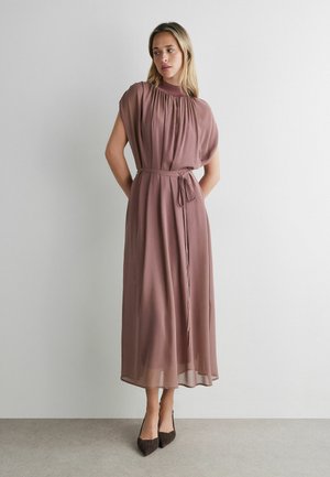 Kobieta w mauve'owej sukience midi bez rękawów z marszczonym dekoltem i wiązaniem w talii, stojąca przy białej ścianie, nosząca ciemne szpiczaste szpilki.