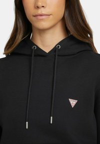 Sudadera negra con capucha y cordón ajustable, con terminales de metal plateado, que presenta un pequeño logotipo triangular rojo y blanco en el lado izquierdo del pecho. Textura de material suave.