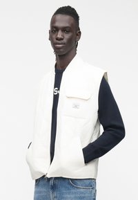 Jaunas vyras, dėvintis baltą siūtinį liemenę ant tamsiai mėlyno megztinio ir mėlynų džinsų, stovintis su rankomis kišenėse balto fono priešakyje.