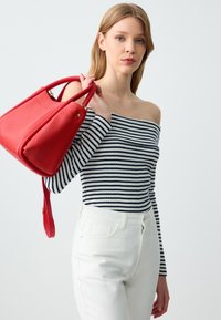 Rote Ledertasche mit strukturiertem Oberflächen, matter Oberfläche und doppelt genähten Griffen, getragen über einem schwarz-weißen gestreiften Off-Shoulder-Oberteil und weißen Jeans.