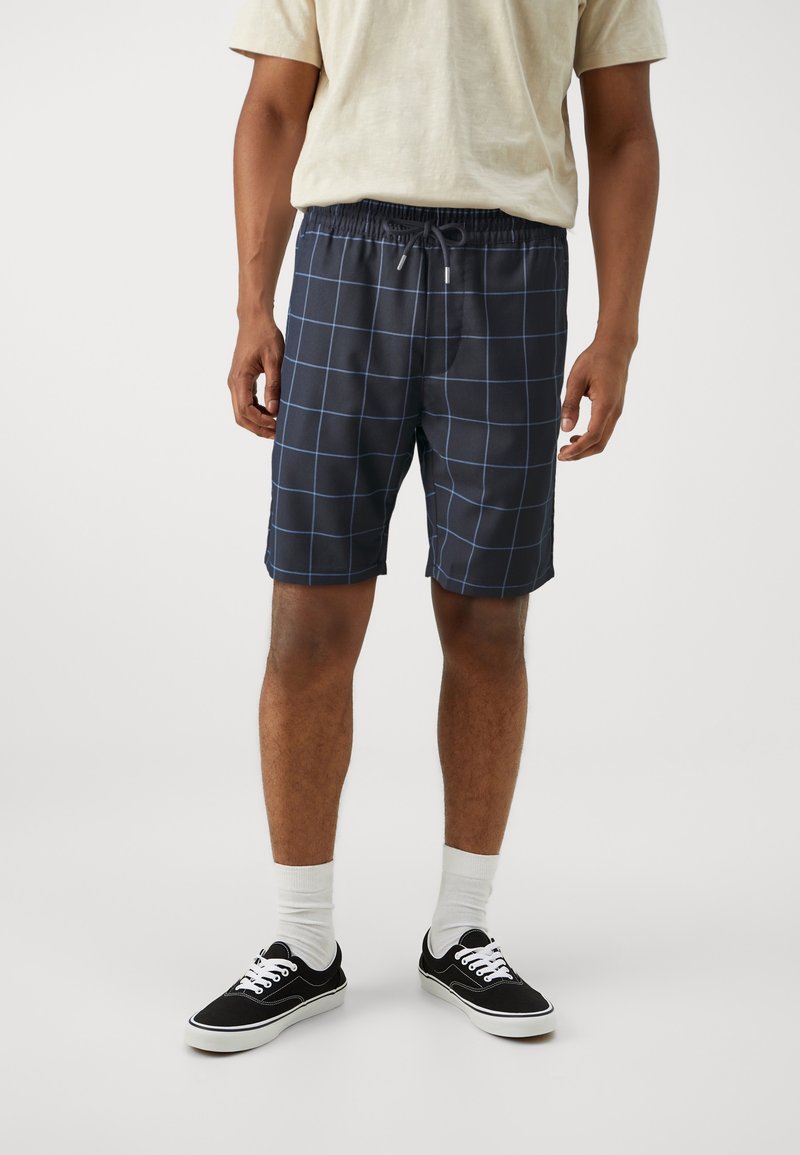 Mørkeblå ternede shorts med elastisk talje og snøre, lavet af et let materiale. Bæret med sorte sneakers og hvide sokker.