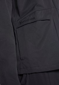 Czarna tkanina kurtki o gładkiej fakturze. Posiada kieszeń na zamek oraz tłoczone logo "Houdini" na górnej części kieszeni.