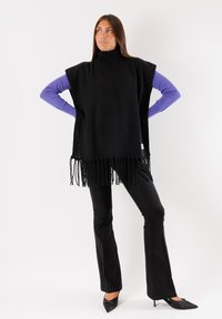 Poncho lavorato a maglia nero con collo alto, rifinito con lunghe frange all'orlo. Indossato sopra una maglietta a maniche lunghe viola e pantaloni neri a zampa.
