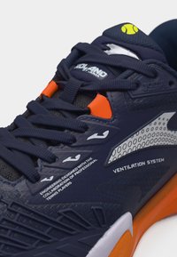 Τένις παπούτσι σε ναυτικό μπλε με πορτοκαλί λεπτομέρειες, μαύρα κορδόνια, σύστημα αερισμού και κείμενο που τονίζει τη συνεργασία με επαγγελματίες παίκτες και τον σχεδιασμό μηχανικής.