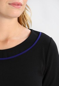 Czarna bluzka z okrągłym dekoltem, wykończona kontrastowym fioletowym obszyciem i gładką fakturą. Zaprojektowana z dopasowaniem na ramionach.