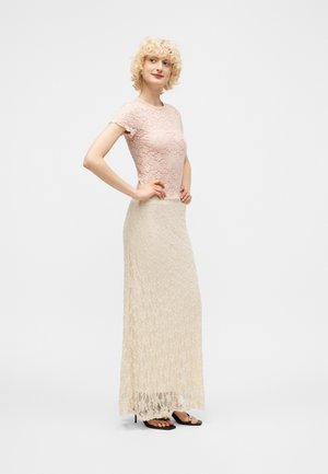 Γυναίκα με σγουρά ξανθά μαλλιά που φοράει ροζ δαντελένιο κοντομάνικο τοπ και μακριά μπεζ δαντελένια φούστα, στέκεται με τα χέρια στη μέση.