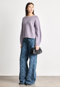 Νεαρή γυναίκα με πουλόβερ πλεκτό λιλά, μπλε παντελόνια φαρδιά με σχέδια, μαύρα παπούτσια, κρατώντας μικρή μαύρη τσάντα χειρός.