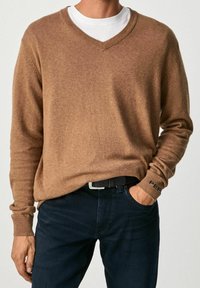 Homme portant un pull marron à col en V par-dessus une chemise blanche, avec un jean bleu foncé et une ceinture noire, une main dans la poche.