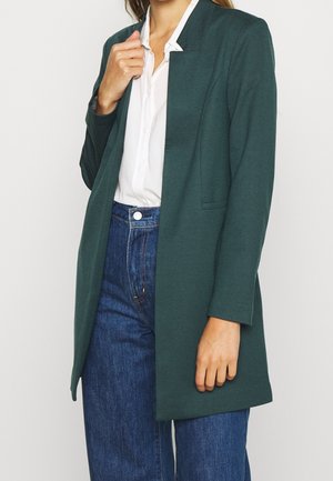 Longue veste blazer vert foncé en tissu lisse. Caractérisée par un design sans col, une ouverture sur le devant et des poches latérales. Assortie à une chemise blanche et un jean en denim.