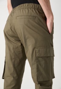 Pantalon cargo vert olive avec une ceinture élastique, des poches latérales et un tissu texturé, avec une poche cargo visible sur la cuisse.