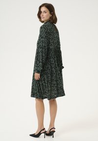 Grönmönstrad klänning med långa ärmar, lös passform, knä-lång fåll och ett nyckelhål bak; kombinerad med svarta slingback-klackar.