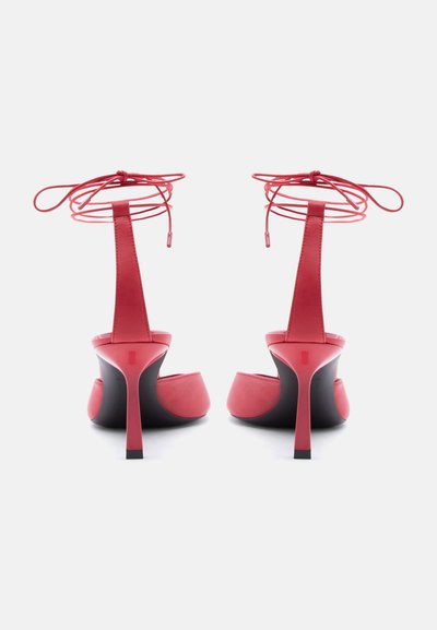 Κόκκινα σανδάλια με ψηλό τακούνι και λουράκια στον αστράγαλο δεμένα σε φιόγκους, φωτογραφημένα από πίσω σε λευκό φόντο.