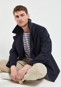 Cappotto di lana blu navy con colletto alto e due bottoni frontali, abbinato a una camicia a righe. Il modello è seduto con le gambe incrociate su uno sfondo neutro.