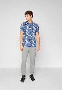Camisa de manga corta con patrón de camuflaje azul y cuello redondo, combinada con pantalones deportivos gris claro y zapatillas deportivas negras.