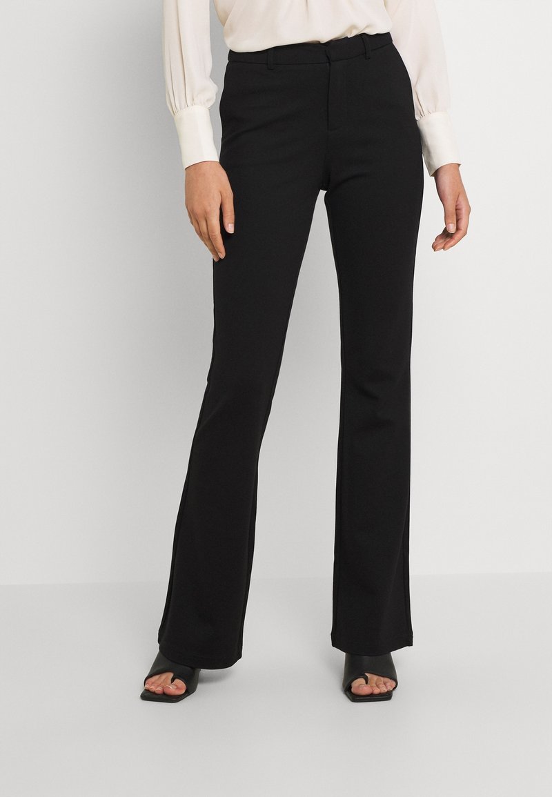 Femme portant un pantalon noir évasé, un chemisier blanc à manches longues, et des chaussures à talons hauts noires à bouts ouverts, debout devant un fond uni.