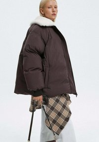 Brązowa kurtka puchowa z białym kołnierzem z sztucznego futra, z zamkiem błyskawicznym i ściągaczami przy rękawach. Nosi ją osoba w zestawie z wzorzystą warstwą i jasnymi spodniami.