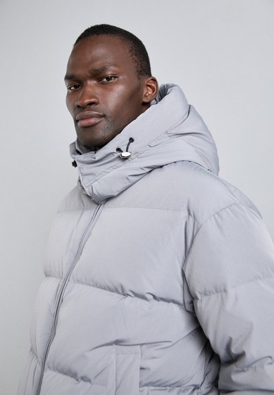 Jachetă puffer de culoare gri deschis, cu închidere prin fermoar, guler înalt și glugă ajustabilă. Prezintă un design matlasat și buzunare laterale. Material neted, rezistent la apă.