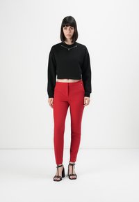 Kvinne som står mot hvit bakgrunn iført svart langermet crop-top, røde smale bukser og svarte høyhælte sandaler.