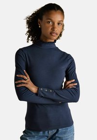 Kortsukkeliste juustega naine, kes kannab tumesinist pikka varrukatoppi volangkrae ja nööpidega mansettidega, käed ristis, seistes ühtlase tausta ees.