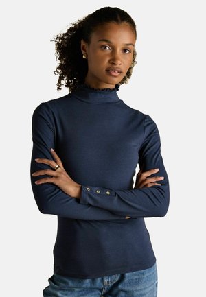Woman with curly hair wearing a navy blue long-sleeve top with a ruffled collar and buttoned cuffs, arms crossed, standing against a plain background.