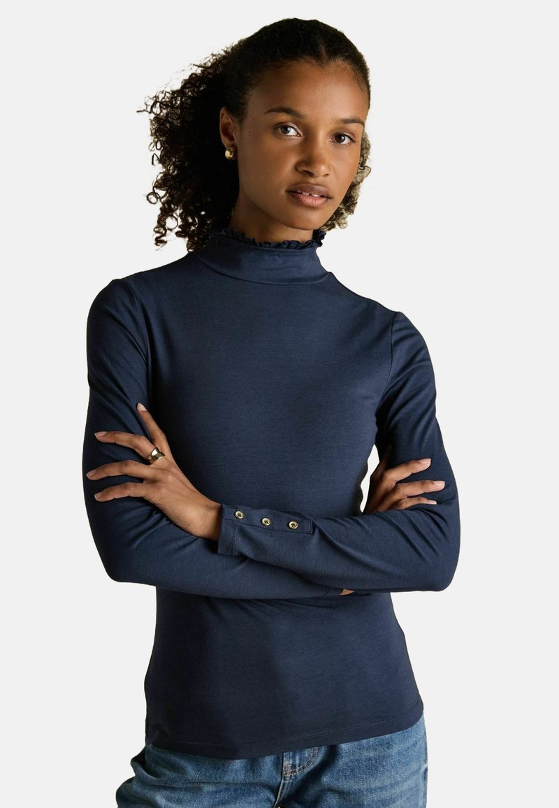 Kortsukkeliste juustega naine, kes kannab tumesinist pikka varrukatoppi volangkrae ja nööpidega mansettidega, käed ristis, seistes ühtlase tausta ees.