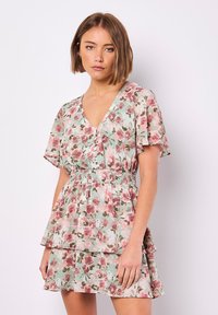 Blumenkleid mit V-Ausschnitt, kurzen flatternden Ärmeln und einem smockierten Bund. Die Farben umfassen mintgrün mit pinken und roten Rosen, sowie einen geschichteten Rock.