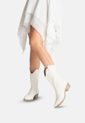 Ben iført en hvid asymmetrisk kjole og hvide cowboystøvler med træhæl på en lys baggrund.