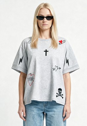 Femme blonde portant des lunettes de soleil noires, un t-shirt gris oversize avec des symboles noirs et rouges, et un jean bleu clair sur un fond uni.