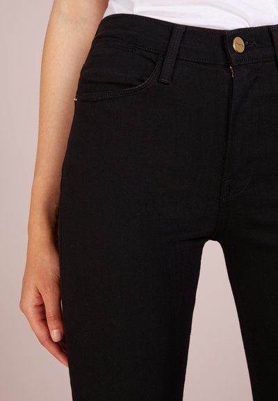 Svarta denimjeans med slim fit, med en gyllene knapp och dragkedja, fem fickor och en mjuk textur. Närbild av midjebandet.