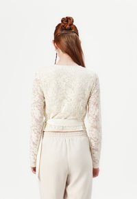 Blonde lace topp med lange ermer og en tettsittende midje, kombinert med kremfargede bukser. Laces har blomstermønstre og en lett glans.