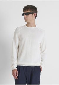 Man met zwarte rechthoekige zonnebril, witte lange mouwen geribbelde trui en donkerblauwe broek met één hand in zijn zak, kijkt naar links.