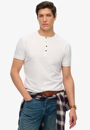 Hvid kortærmet T-shirt med henley-desgin og fire sorte knapper. Bæres med blå jeans og en ternet skjorte bundet omkring taljen.
