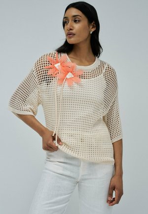 Top de punto de malla crema con un ajuste holgado, adornado con un grupo decorativo de flores de color coral y mangas cortas, combinado con pantalones blancos.