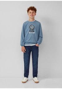 Ragazzo con capelli rossi e ricci sorride, indossando una felpa blu con grafica di faccina sorridente, jeans scuri e sneakers bianche, in piedi contro un muro bianco.