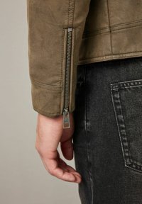 Chaqueta de cuero verde oliva con detalles en cremallera plateada en las mangas, combinada con jeans de mezclilla oscura que tienen un bolsillo trasero. Acabado texturizado visible.