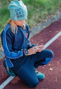 Barnet bär en ljusblå mössa med texten "COOL AND KIND", en marinblå sweatshirt med blå detaljer och mörka byxor. Har på sig en smartklocka.