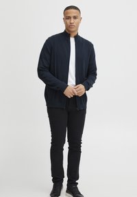 Marineblå glidelås-kardigan laget av mykt strikket stoff, med høy krage, ribbestrikkede mansjetter og nederkant, kombinert med svarte slim-fit bukser og svarte sneakers.