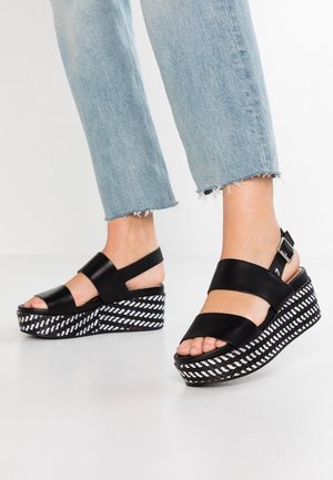 Czarne skórzane sandały na platformie z szerokimi paskami, z wzorzystą czarno-białą podeszwą słupkową i regulowanym paskiem w okolicy kostki.