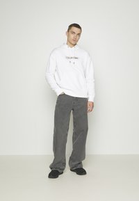 Felpa bianca con una tasca frontale e lacci, con un logo. Abbinata a jeans grigi a taglio largo e scarpe nere. Sfondo semplice.