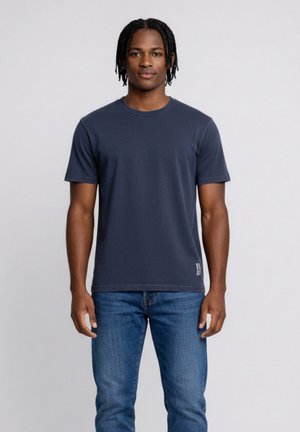 Giovane uomo con capelli corti intrecciati che indossa una maglietta blu scuro a girocollo e jeans blu, in piedi contro uno sfondo chiaro uniforme.