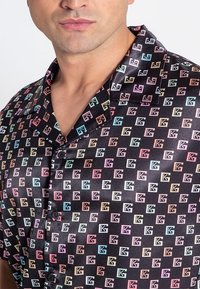 Schwarzes T-Shirt mit kurzen Ärmeln, das ein wiederholtes geometrisches Logomuster in Pastellfarben zeigt. Glatte, satinartige Textur mit Kragen und Knöpfen.