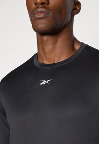Schwarzes Sportshirt aus glattem, leichtem Material. Mit rundem Ausschnitt und kleinem weißen Logo auf der Brust.