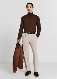 Brauner Rollkragenpullover, beige maßgeschneiderte Hose und eine braune Jacke mit Lammfellfutter, die am Arm gehalten wird; ein schwarzer Gürtel und dunkle Schuhe runden das Outfit ab.