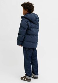 Tumši zila pūdera jaka ar kapuci, ar šūtu dizainu un brīvi pieguļošu piegriezumu, kombinēta ar džinsa biksēm un brūnām sporta kurpēm ar baltām detaļām.