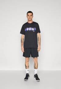Sort grafisk t-shirt med et farverigt "ARDA" print, parret med sorte shorts, hvide sokker og sorte sportssko. Ensfarvet baggrund.